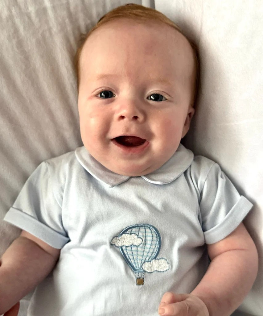 Baby wearing a onesie with a hot air balloon design, lying on a white surface.