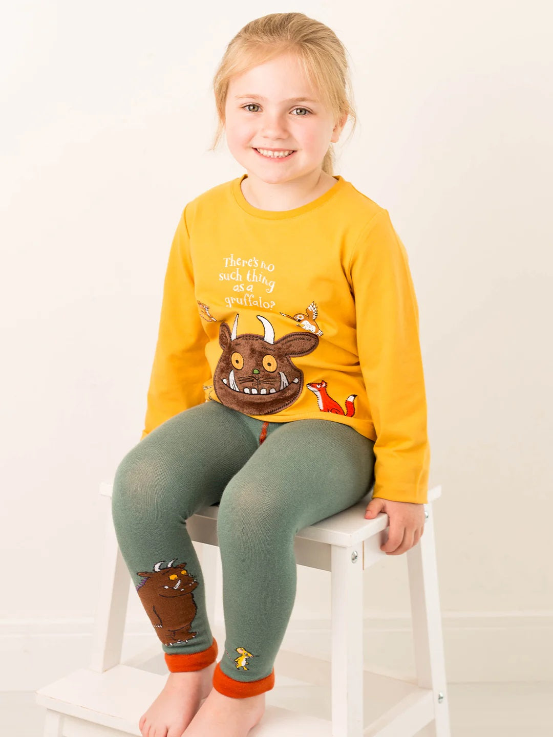 Child wearing a yellow shirt with a cartoon character and green pants sitting on a white stool.