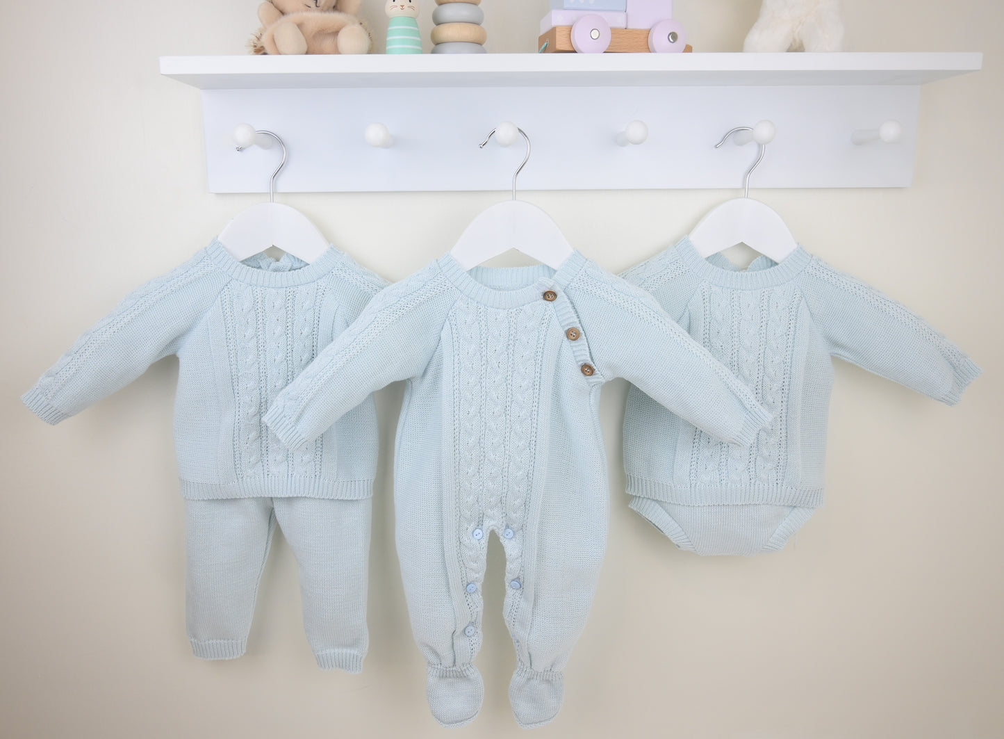 Light blue baby outfits hanging on a white coat rack against a beige wall.