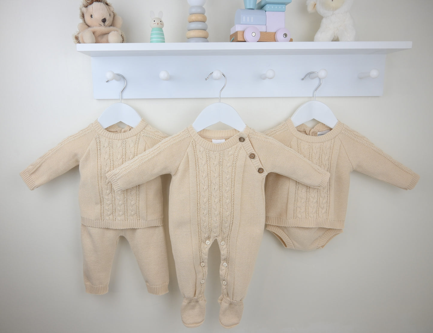 Three beige baby outfits hanging on a white wall with a shelf above containing toys.
