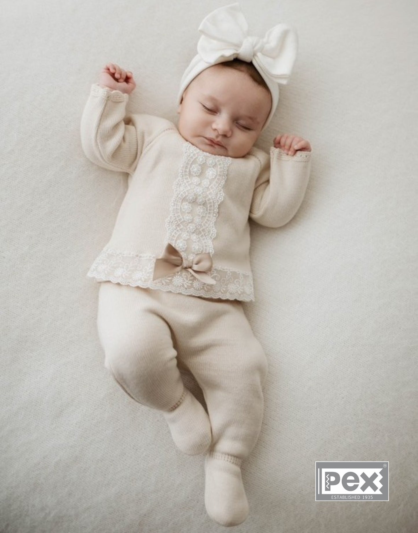 Baby in a natural outfit with a bow and headband, lying on a light gray surface.