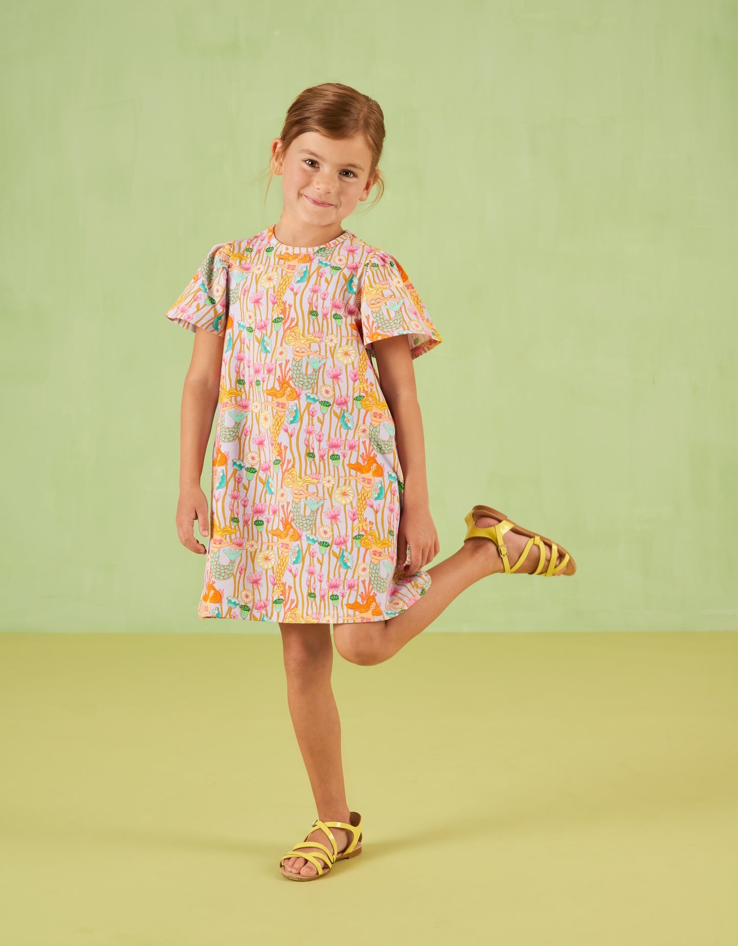 Young girl wearing a colorful Oilily dress with a pattern, standing on a light green background
