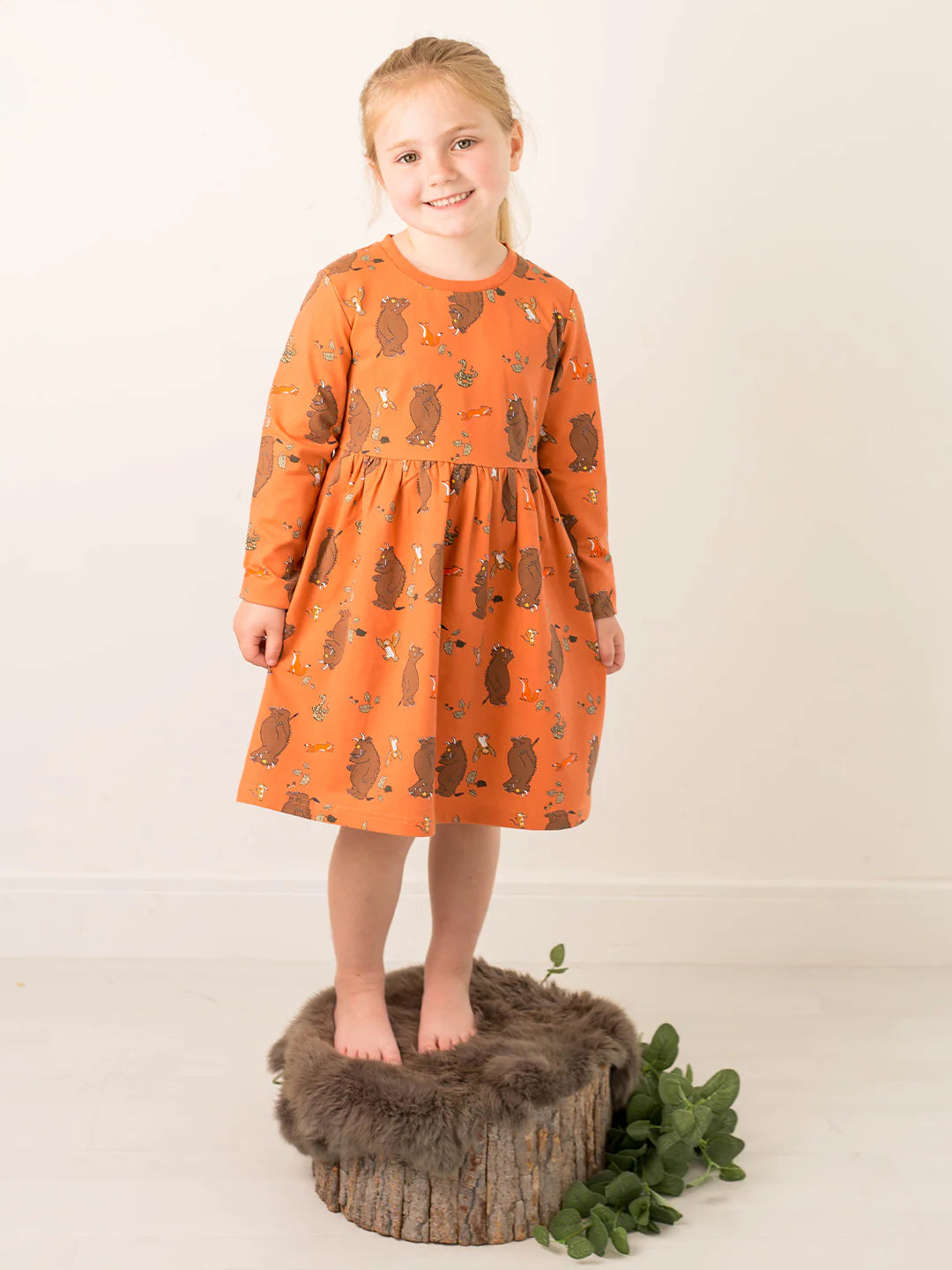 Young girl wearing an orange dress with a Gruffalo pattern, standing on a wooden block with a fur rug and greenery.