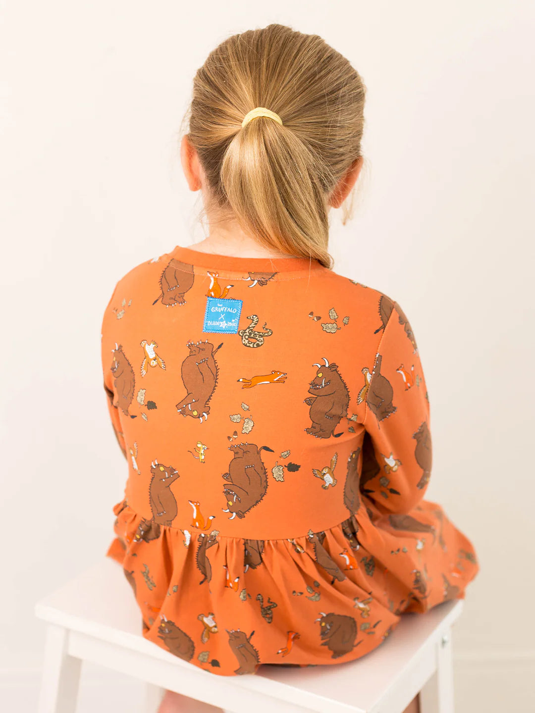 Child wearing an orange dress with Gruffalo pattern sitting on a white chair.