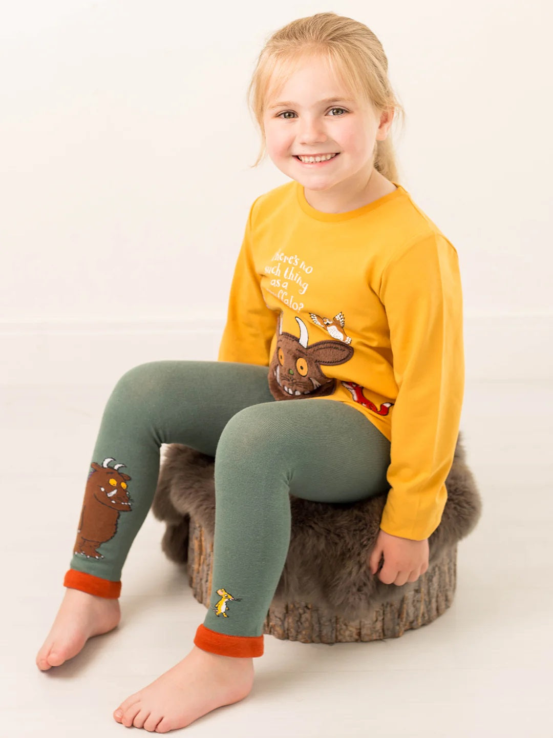Child wearing a yellow long-sleeve shirt and green leggings with Gruffalo designs, sitting on a wooden stool.