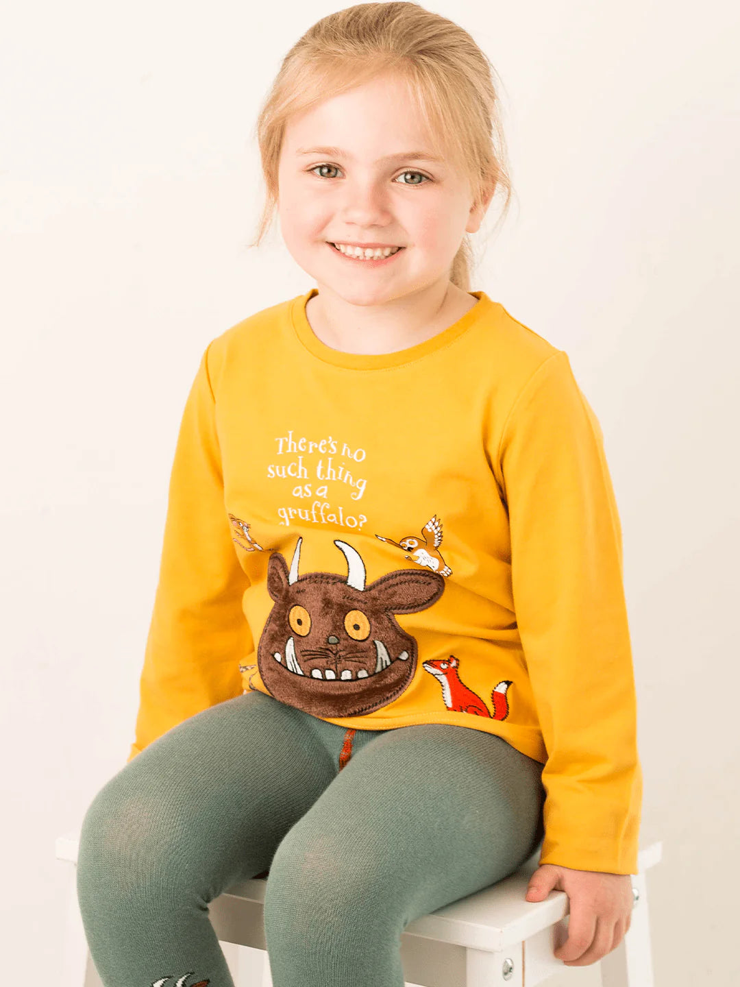 Child wearing a yellow long-sleeve shirt with a gruffalo character and text, sitting on a white stool.