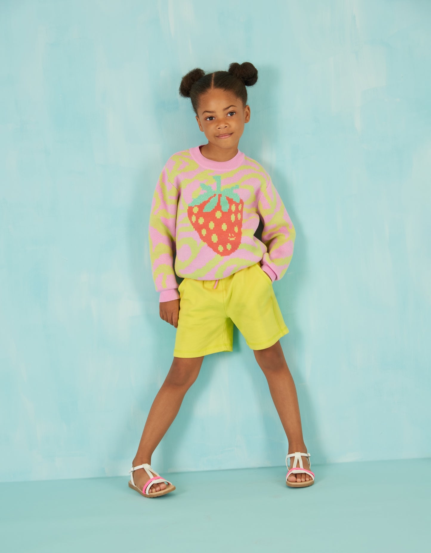 Child wearing a colorful strawberry sweater and yellow shorts against a light blue background