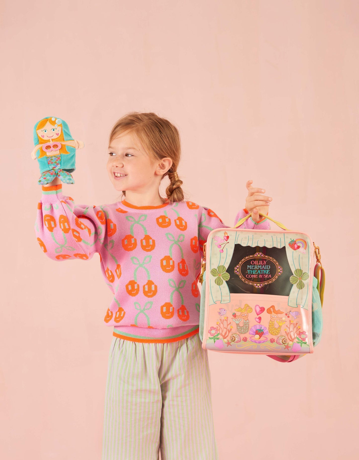 Child wearing a pink sweater with cherry design, holding Oilily mermaid theatre bag and a toy on a pink background