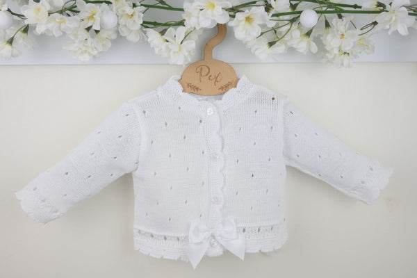 White baby cardigan with a bow on a light background with floral decorations.
