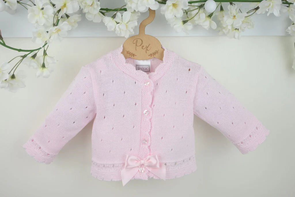 Pink baby cardigan with a bow on a hanger against a light background with flowers.