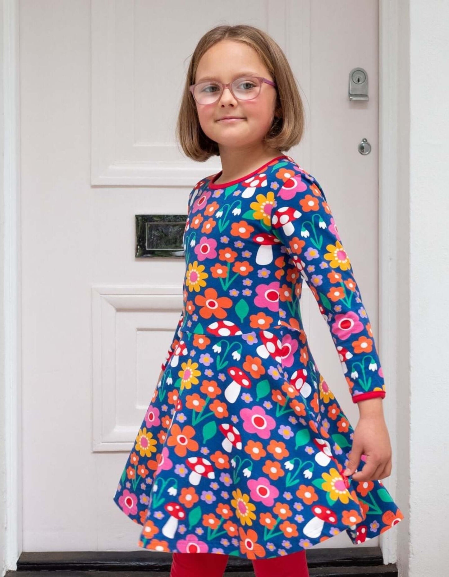 Young girl wearing a colorful floral Toby Tiger dress standing in front of a white door.
