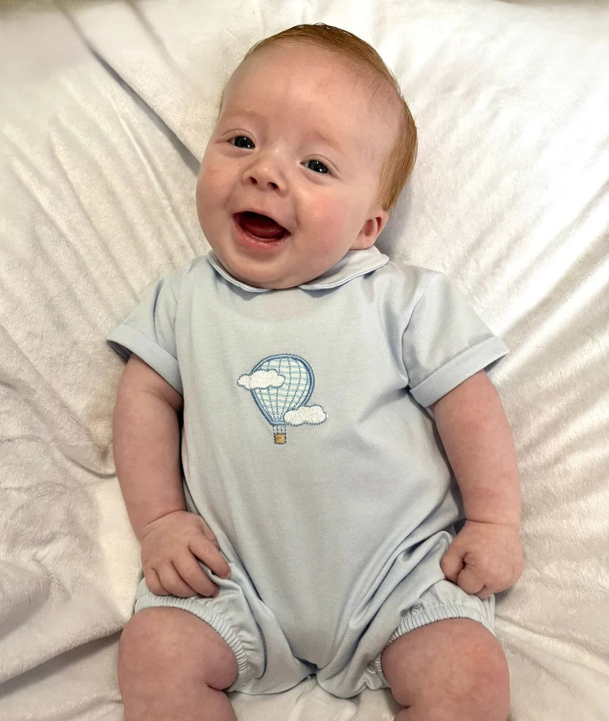 Baby wearing a light blue onesie with a hot air balloon design on a white blanket.