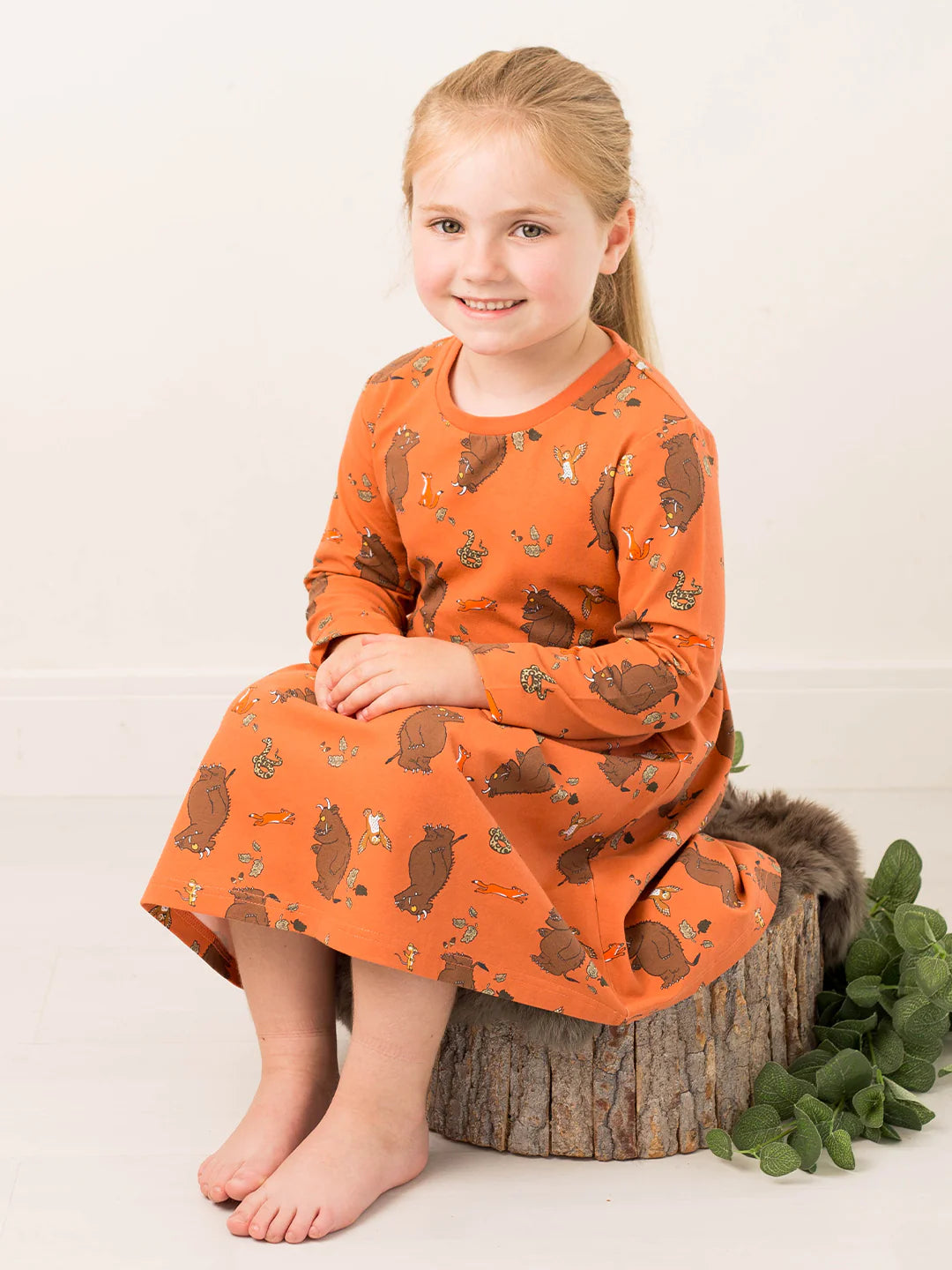 Young girl wearing an orange dress with gruffalo pattern sitting on a log.
