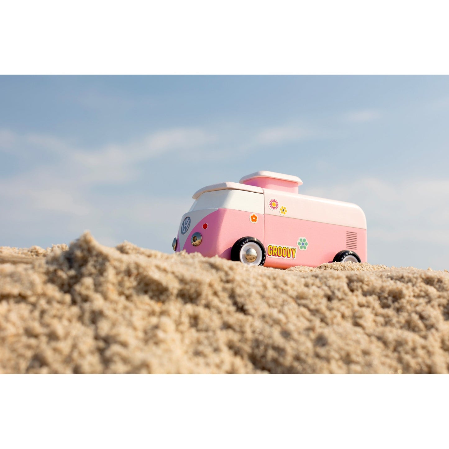 Pink and white vintage-style camper van on a sandy beach with a clear blue sky.
