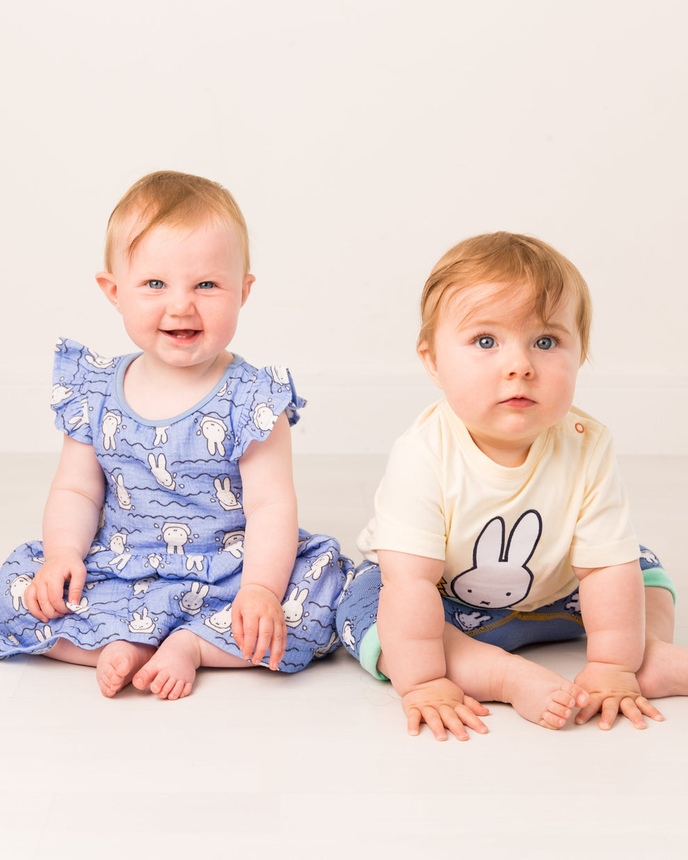 Two babies sitting on a white floor wearing Miffy patterned outfits.