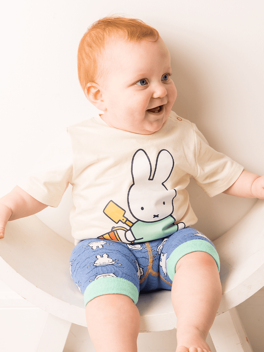 Baby wearing a white shirt with a Miffy cartoon character and blue shorts sitting on a white surface.