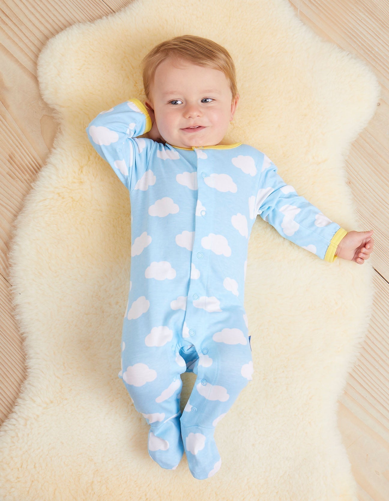 Baby wearing a blue babygrow with white cloud patterns, lying on a soft beige surface.