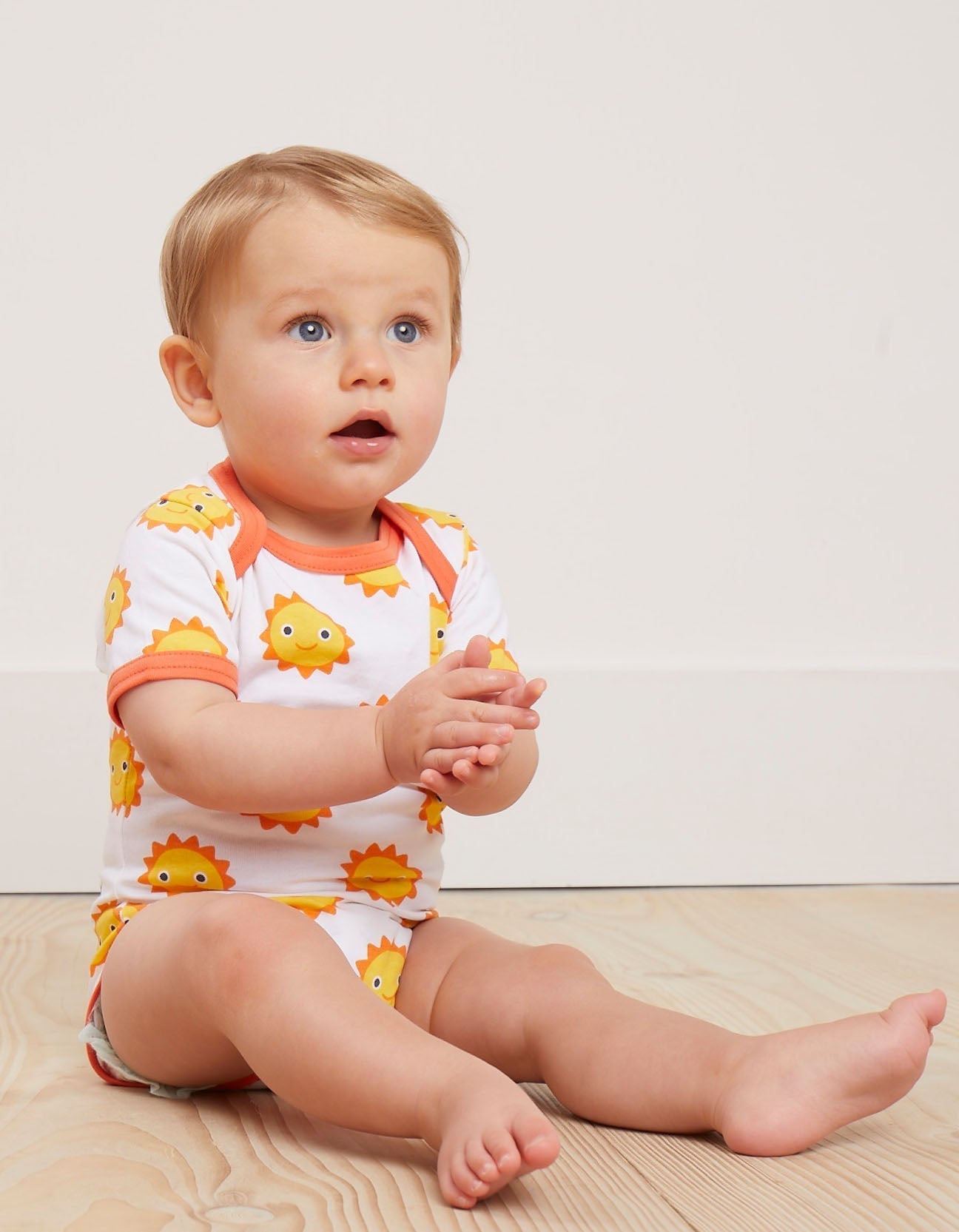Baby wearing a sun-themed onesie sitting on a wooden floor.