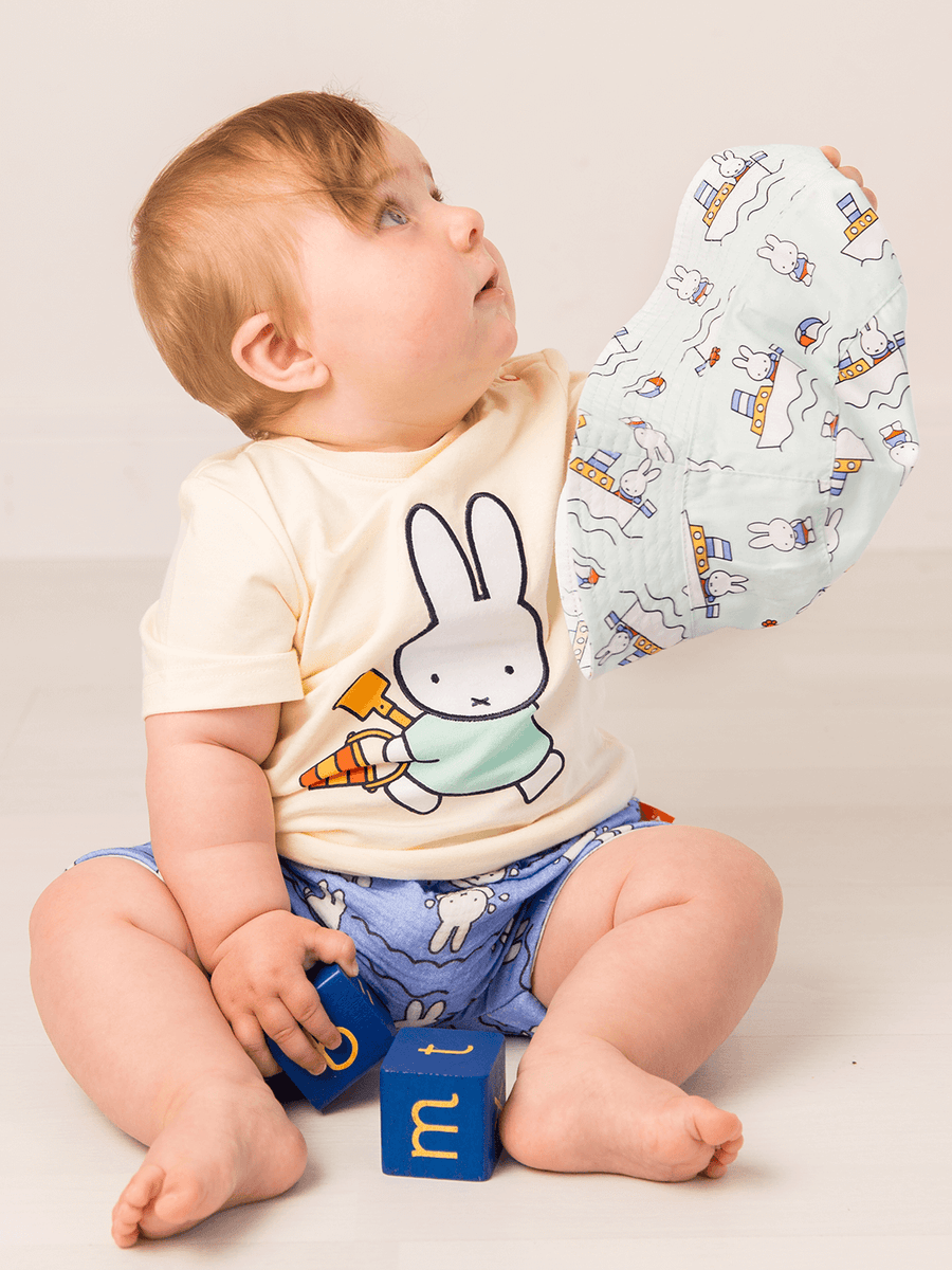 Baby wearing a tshirt with a miffy cartoon character and patterned pants, holding a baby hat.