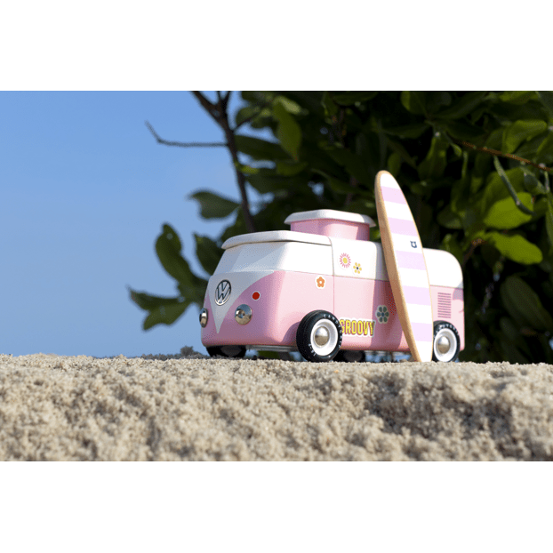 Pink toy van with a surfboard on a sandy surface with greenery in the background