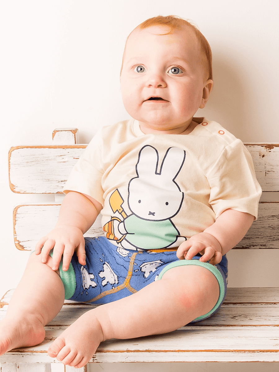 Baby wearing a miffy tshirt with a cartoon character sitting on a wooden bench.