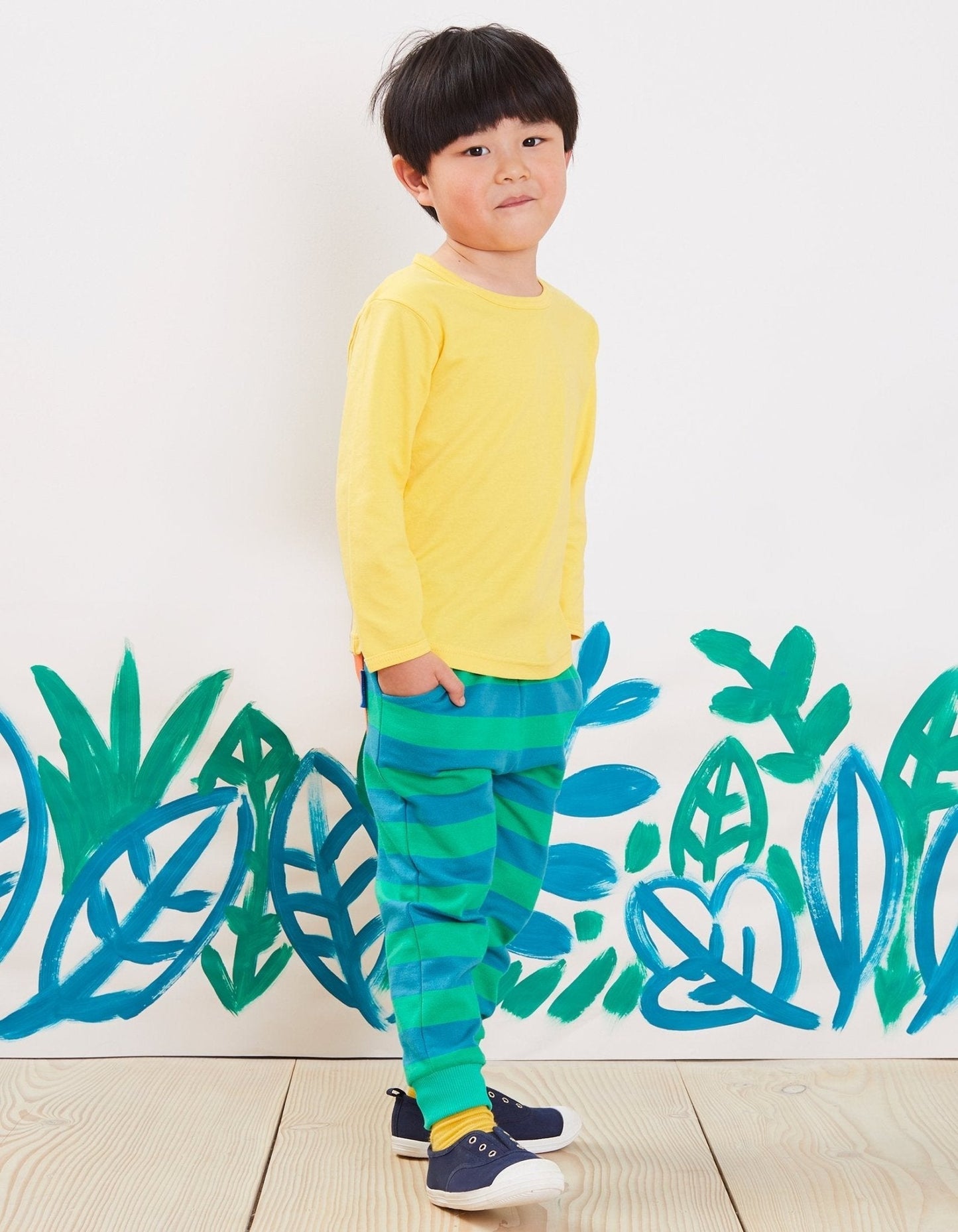 Child wearing a yellow shirt and green striped joggers standing in front of a white wall with blue and green leaf patterns.
