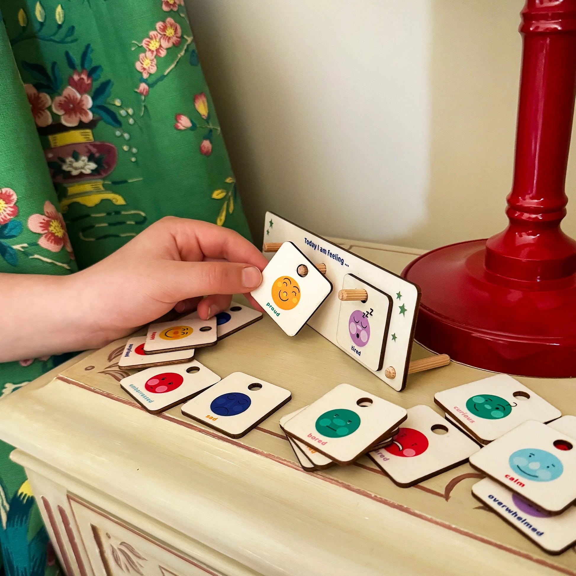 Emotions feeling chart with cards on a wooden surface, next to a red lamp and floral curtain.