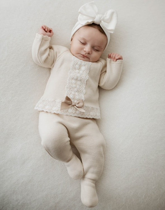 Baby in a natural outfit with lace details and a bow, lying on a light gray surface.