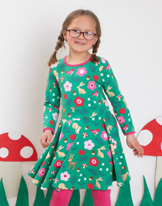 Young girl wearing a green dress with floral and animal patterns, standing against a white background.