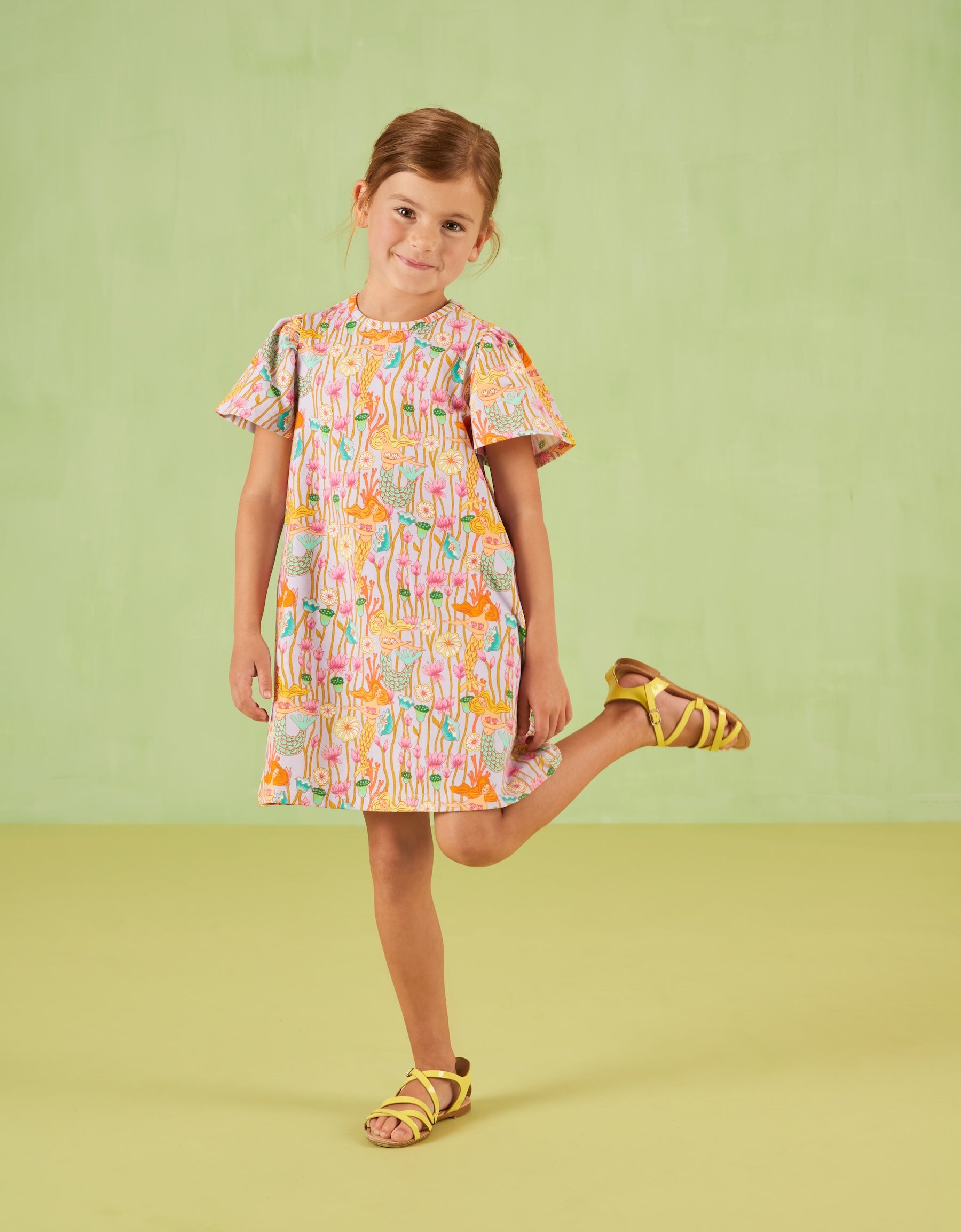 Young girl wearing a colorful Oilily dress with a pattern, standing on a light green background