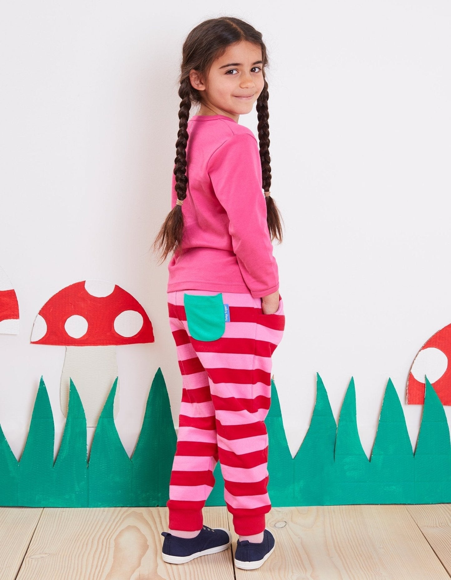 Young girl wearing a pink top and red and pink striped joggers standing in front of a mushroom and grass backdrop.