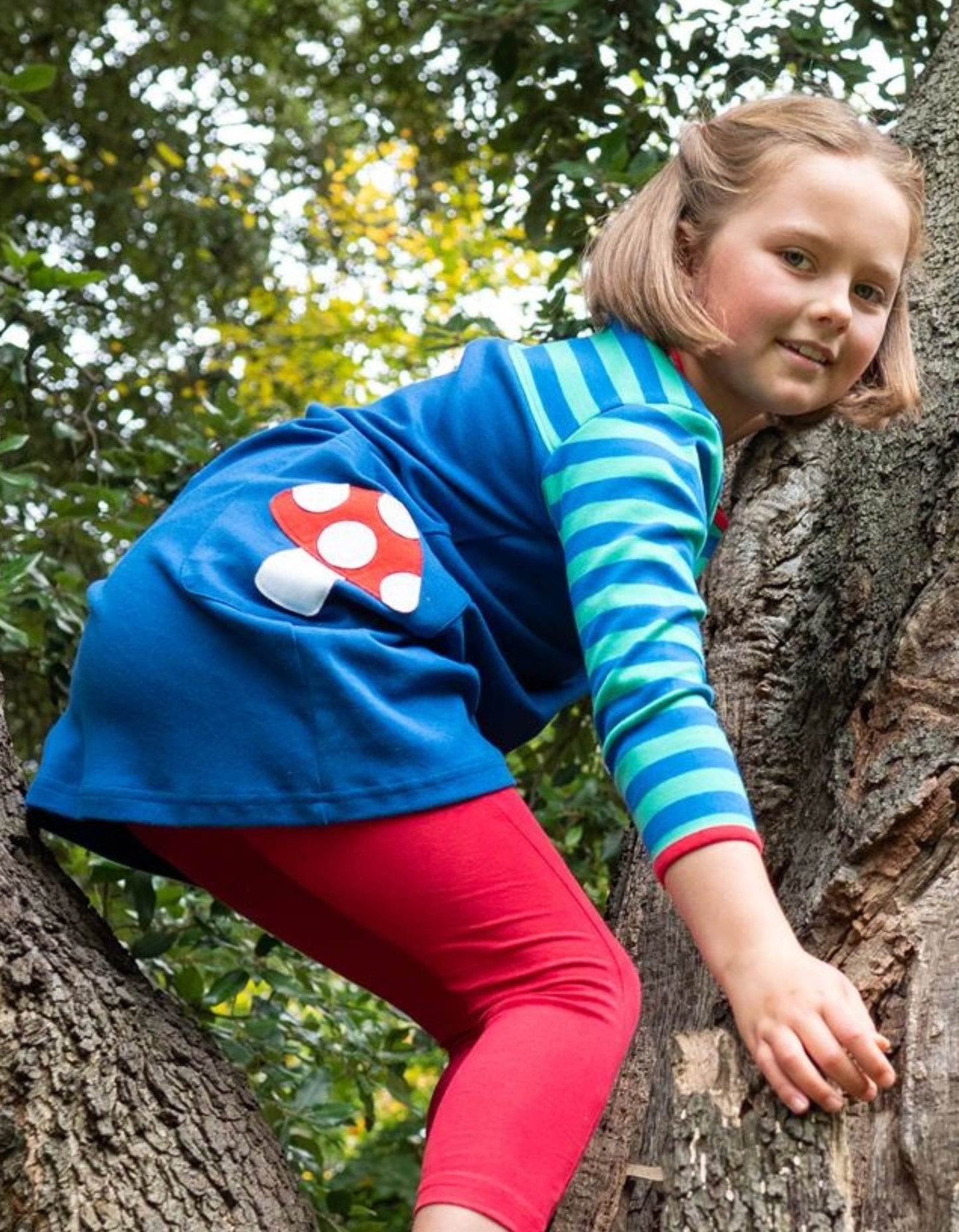 Child climbing a tree wearing a blue dress with a red and white mushroom design.