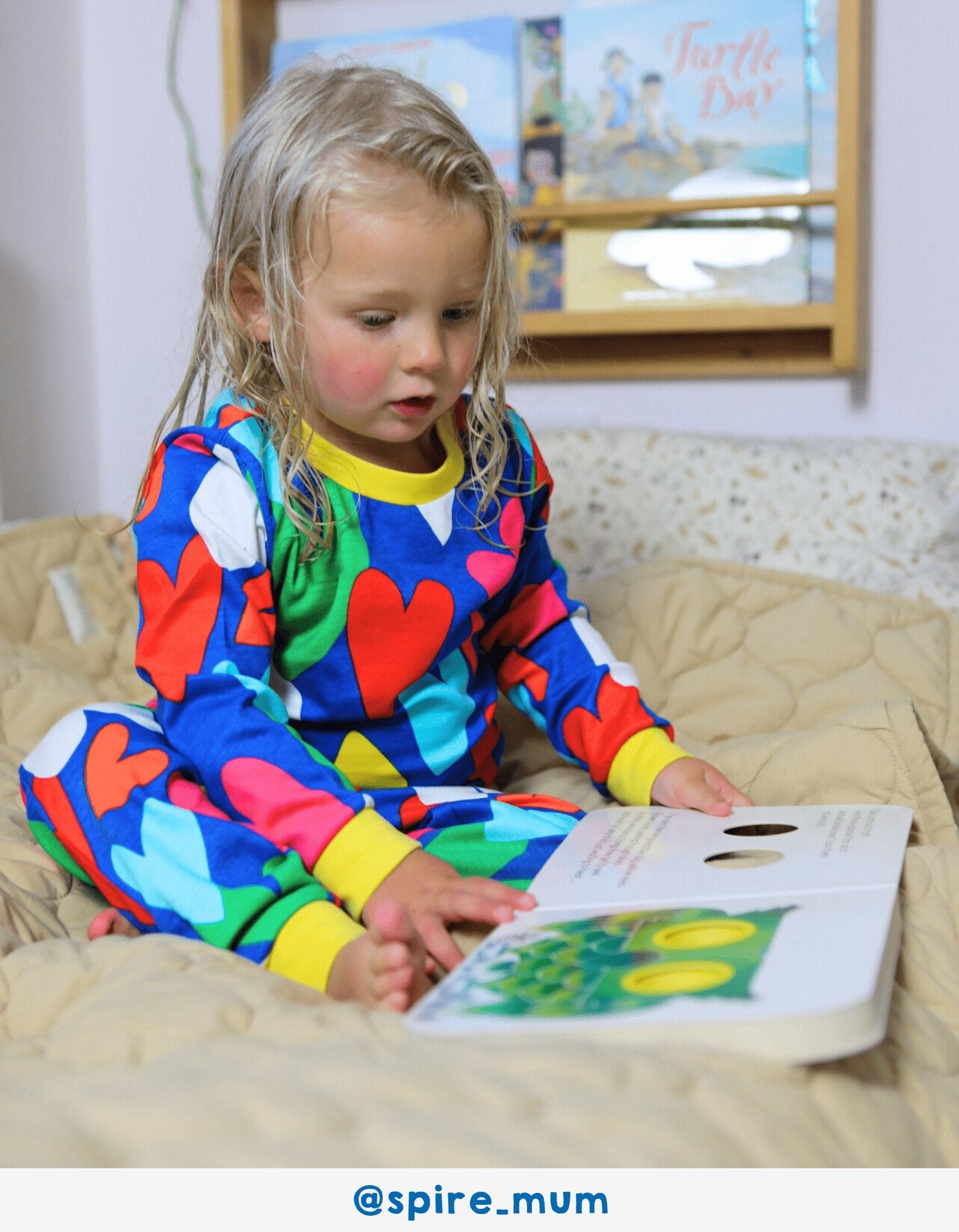 Child in colourful pyjamas reading a book on a bed with a framed picture in the background.