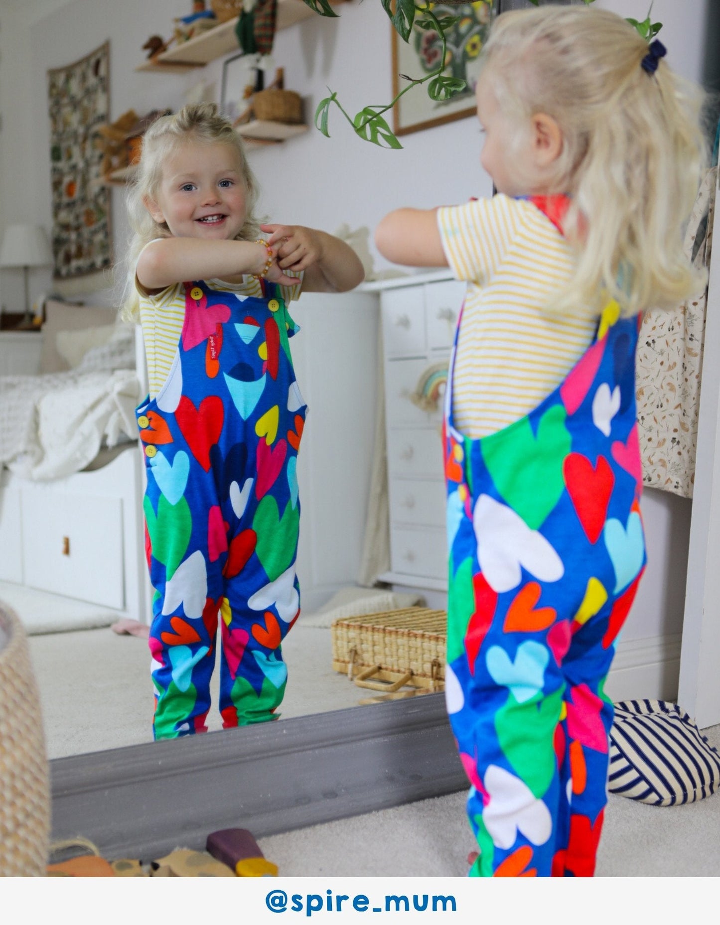 Child wearing colourful heart-patterned dungarees standing in front of a mirror.