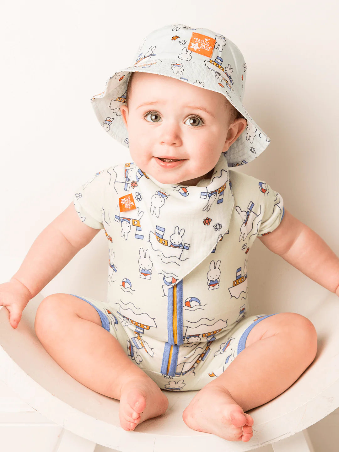 Baby wearing a Miffy patterned romper, bib, and hat sitting in a white chair.