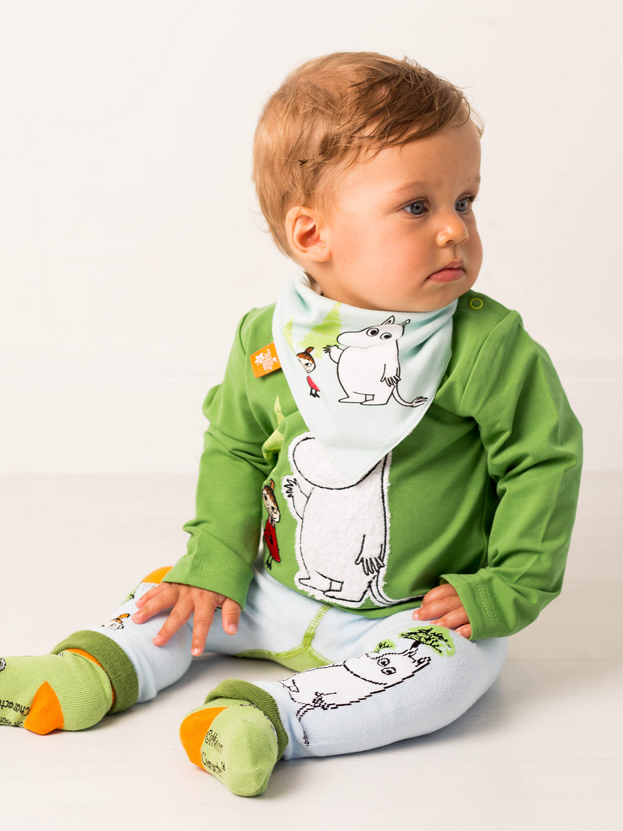 Baby wearing a green top, blue leggings  with Moomin designs, and a matching bib on a white background.