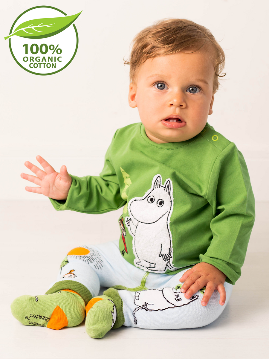 Baby wearing a green shirt with a Moomin cartoon character, on a white background with a 100% organic cotton logo.