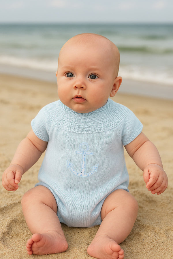 Baby in a light blue onesie with an anchor design sitting on a sandy beach.