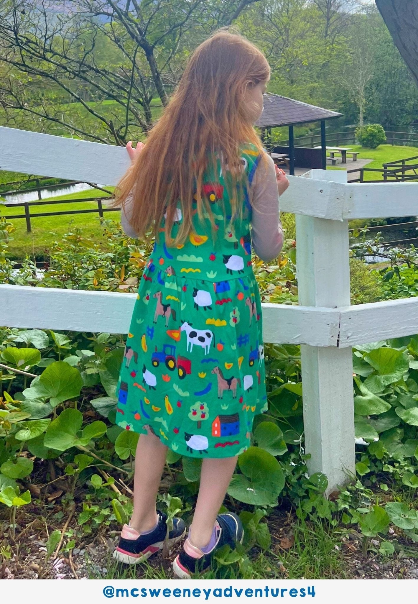 Child wearing a green dress with farm animal prints standing near a white fence outdoors.