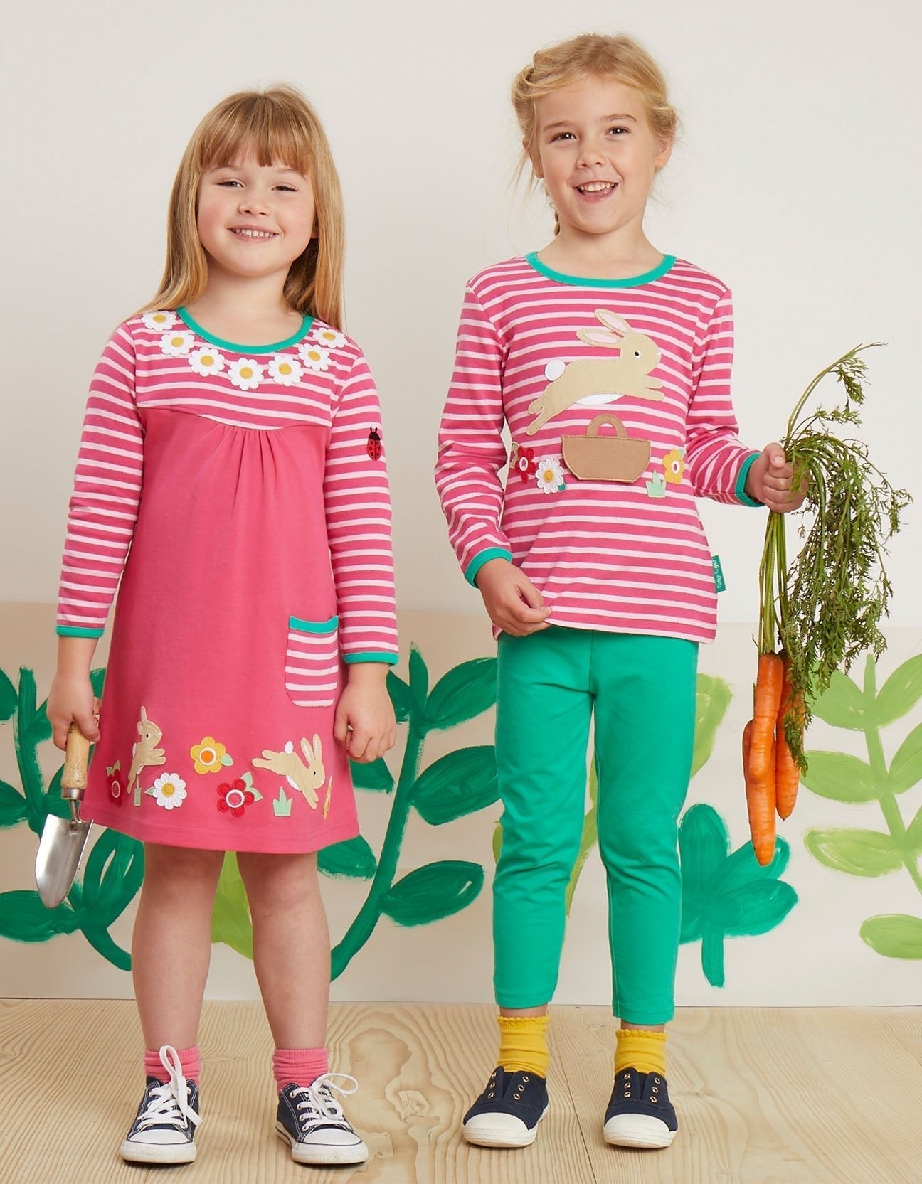 Two children wearing pink and green leaping bunny outfits by Toby Tiger against a leafy green background.