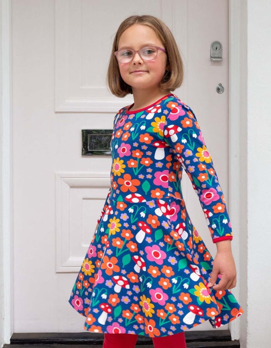 Young girl wearing a colorful floral Toby Tiger dress standing in front of a white door.