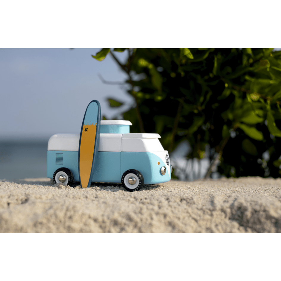 Toy van with a surfboard on a sandy surface with greenery in the background