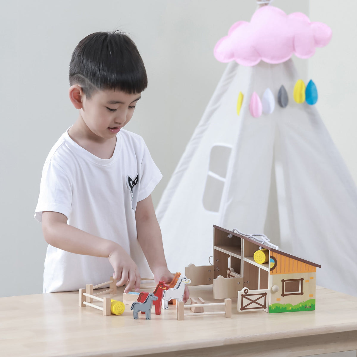 Young child playing imaginatively with an open foldable wooden toy stable and farmyard playset.
